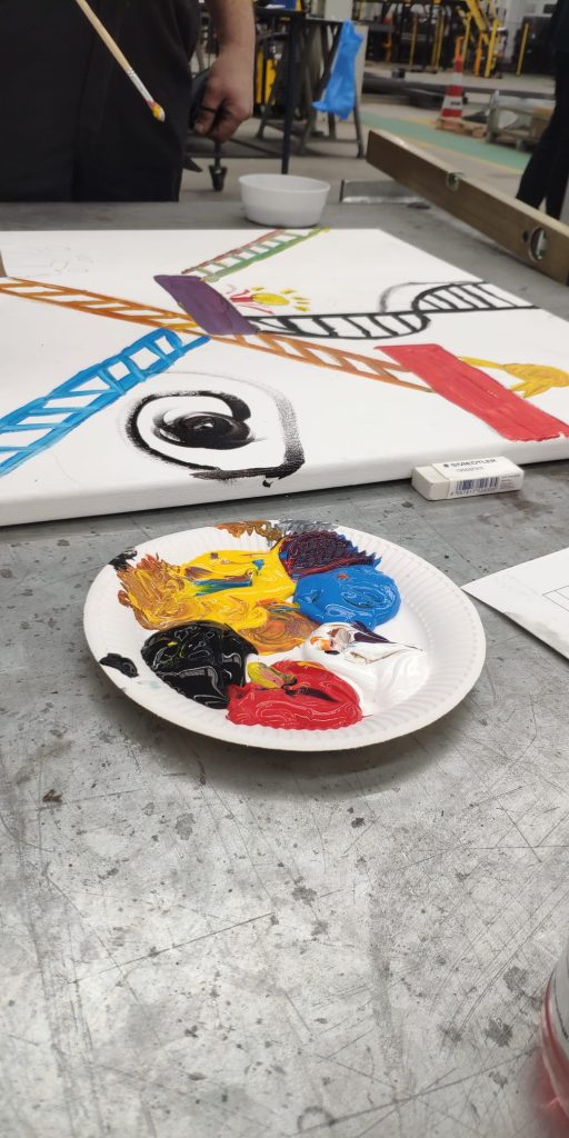 Sfeerbeeld van een workshop. te zien is een bord met verf een schilderij en iemand op de achtergrond die schildert.